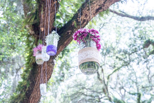 Hanging glass jars with flowers and lace decor on a tree outdoors.