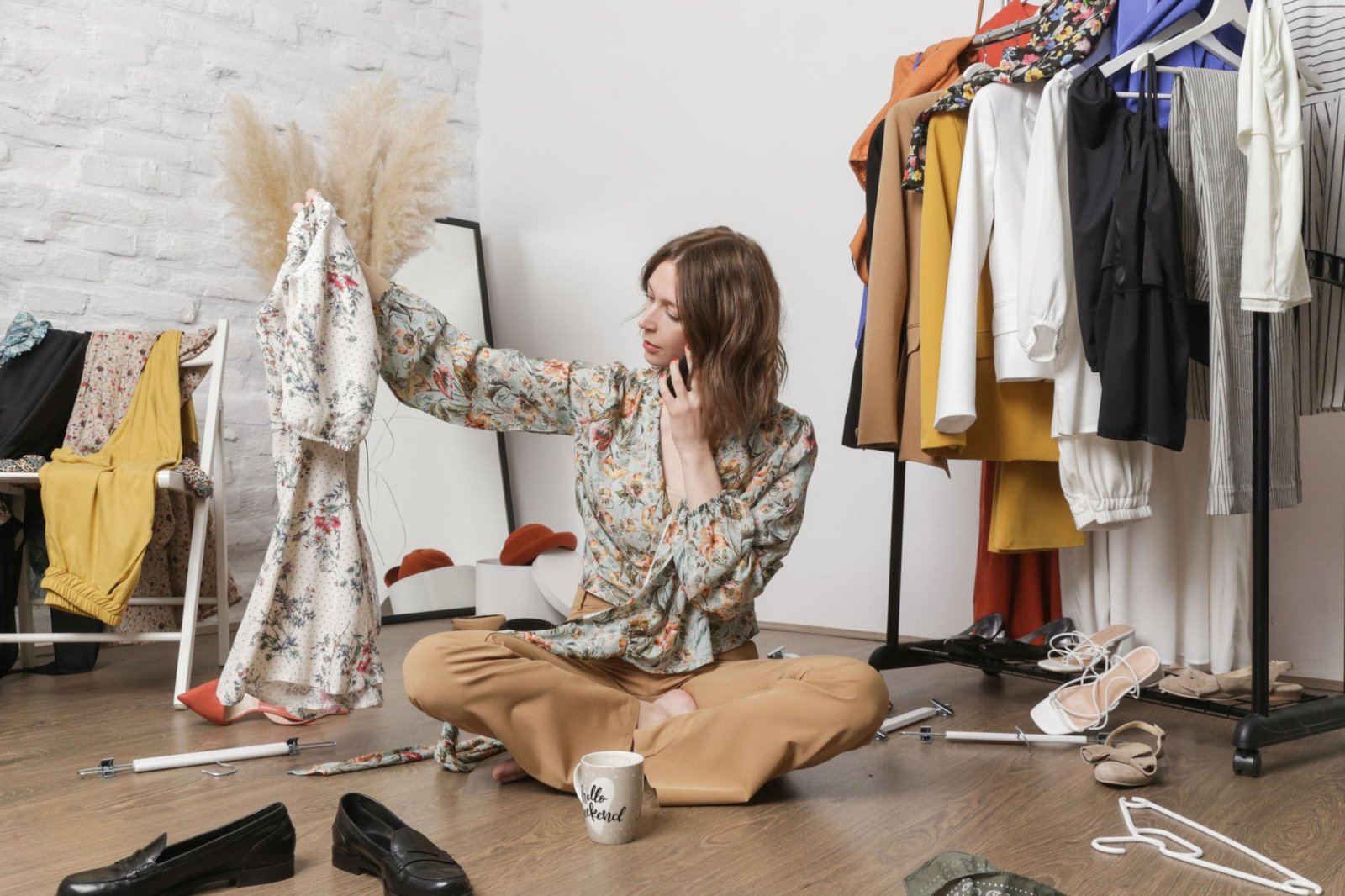 A woman sits cross-legged on a wooden floor surrounded by scattered shoes, hangers, and clothes. She is holding up a floral dress while talking on a smartphone, looking frustrated with her disorganized clothing rack in a white brick room.