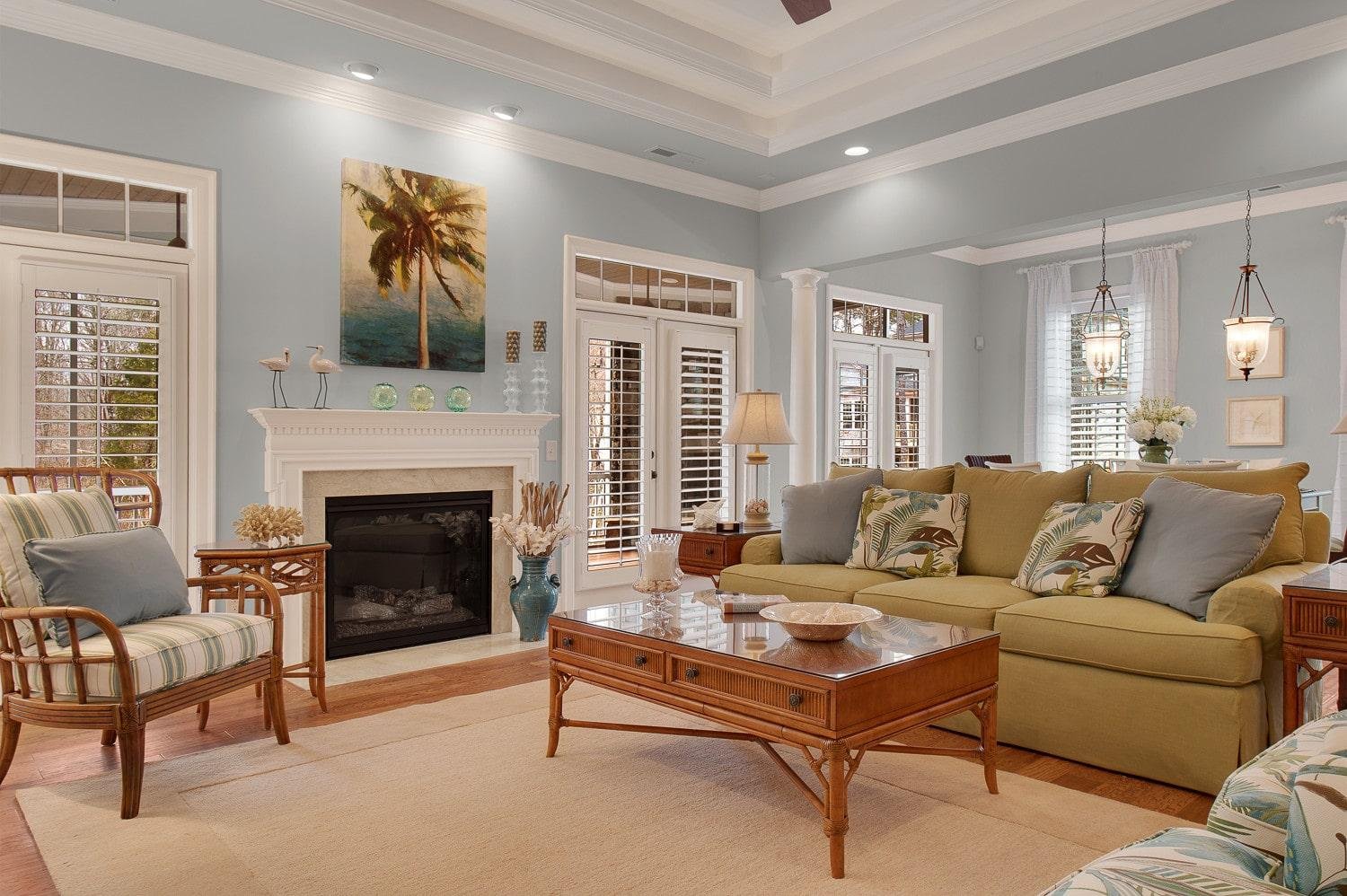 Light blue living room with coastal-style decor, featuring a soft blue-gray wall color, white fireplace, wooden furniture, and neutral sofa with tropical accent pillows.