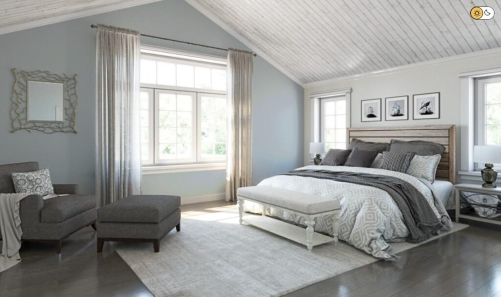 A spacious, bright bedroom featuring a vaulted white wood-plank ceiling and light blue walls. A large bed with a rustic wooden headboard and gray patterned bedding sits on a cream area rug, accompanied by a matching bench, a gray armchair with an ottoman, and sheer curtains framing a large window.