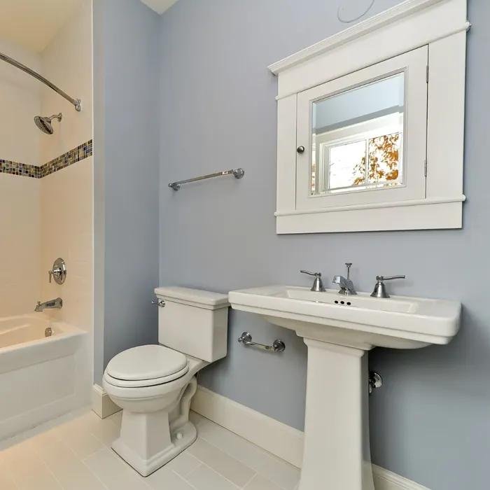 Light blue bathroom with a pedestal sink, white toilet, bathtub with tiled accent strip, and a wall-mounted mirror.