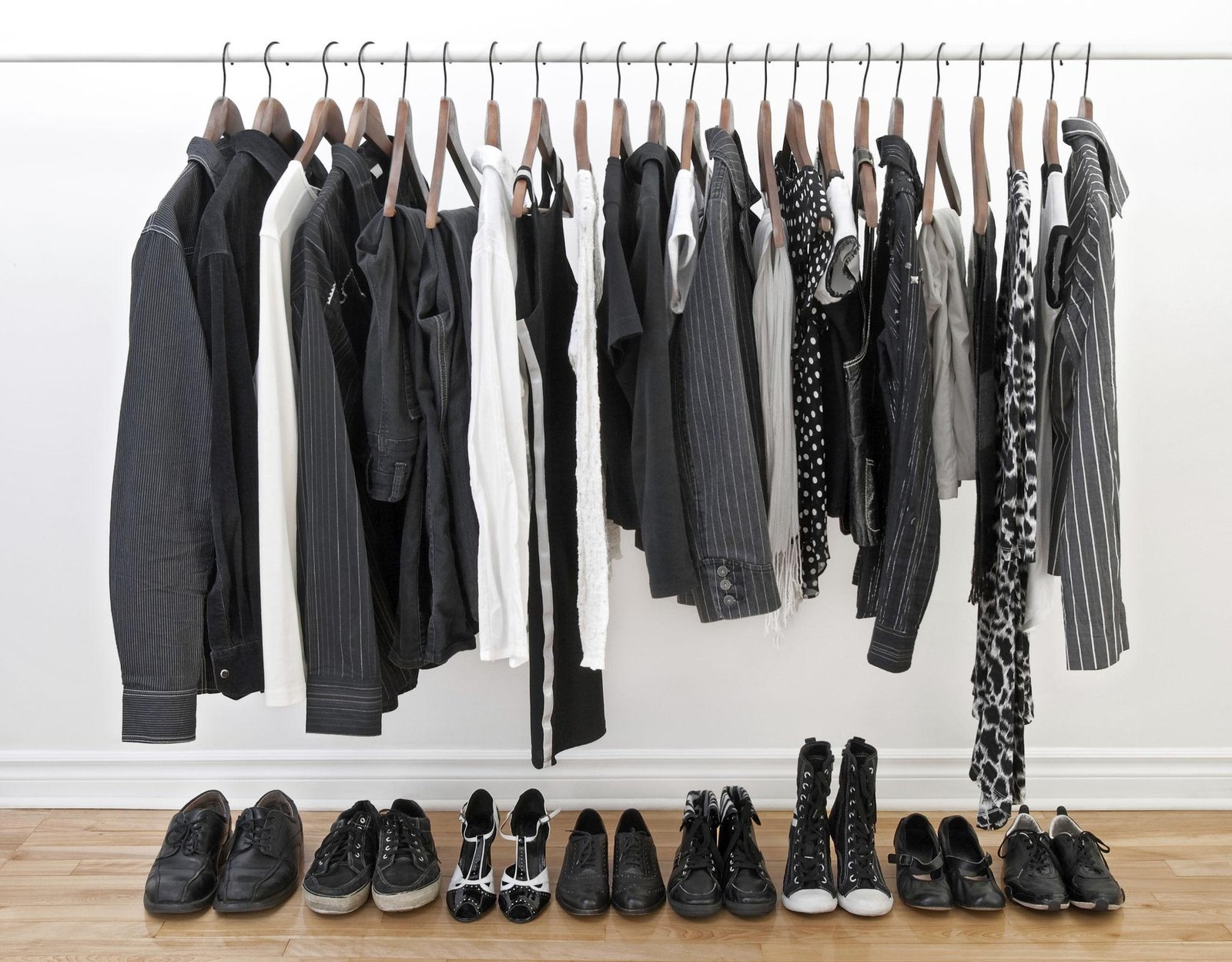 A white clothing rack against a plain white wall holds a curated collection of black, white, and striped garments on wooden hangers. Below the rack, several pairs of black shoes, including sneakers, heels, and boots, are neatly lined up on a light wood floor.
