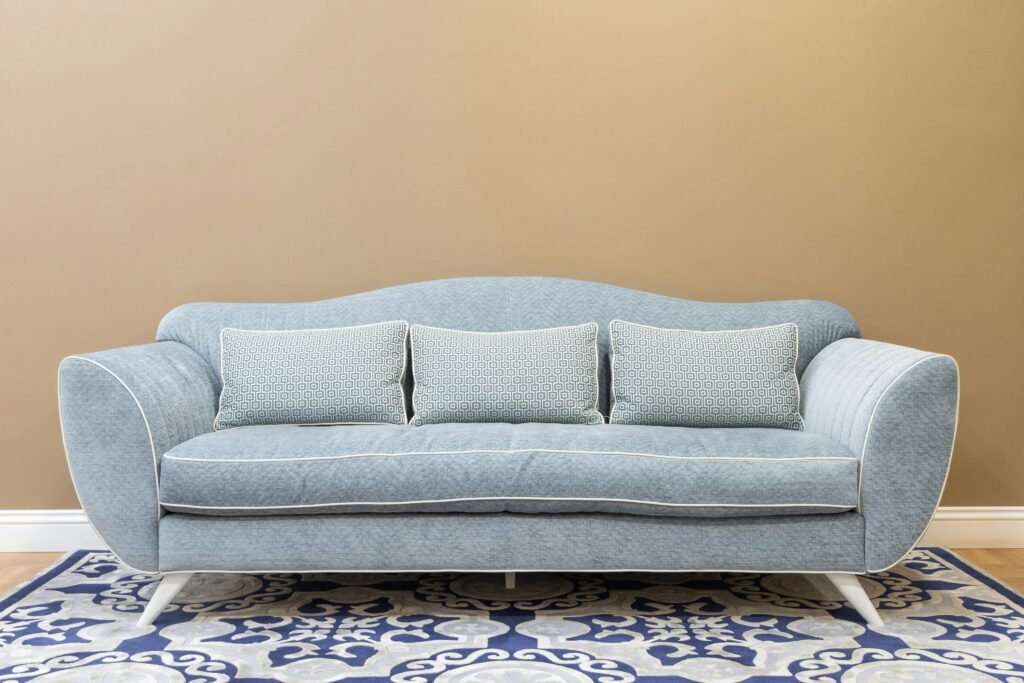 A contemporary light blue fabric sofa with a curved backrest and three matching patterned throw pillows is centered in front of a warm tan wall, resting on a blue and white geometric rug.