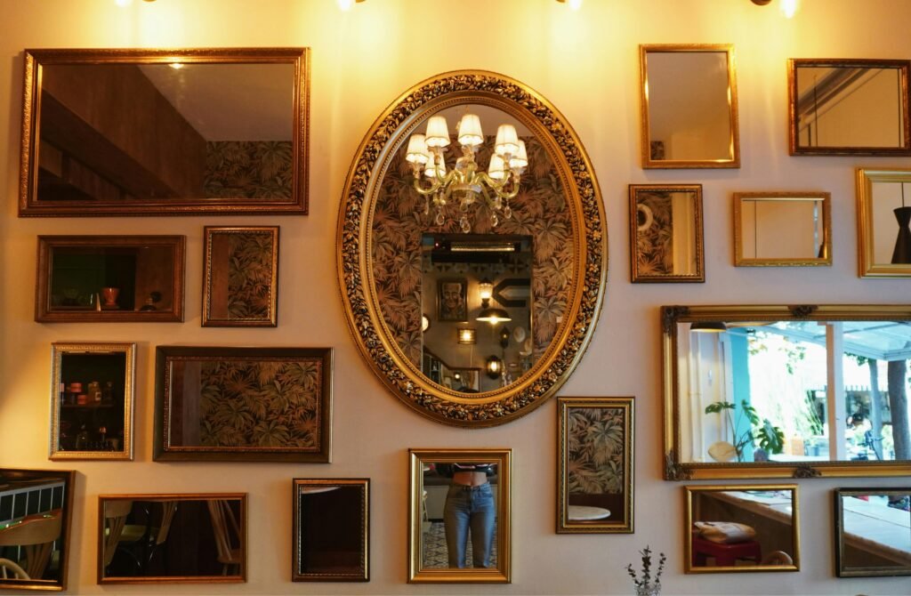 A collection of various antique-style mirrors with ornate gold frames arranged as a gallery wall, featuring a central oval mirror reflecting a classic lit chandelier and tropical palm leaf wallpaper.